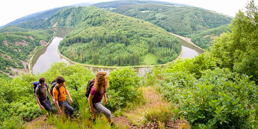 Drei Personen Wandern auf einen Hügel hoch, im Hintergrund fließt ein Fluss